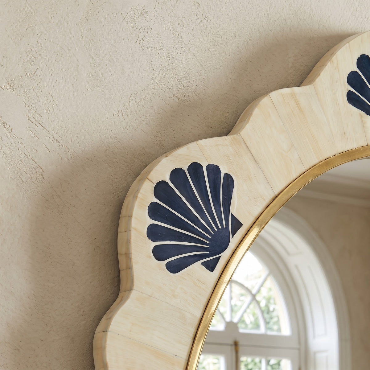 A close-up of a decorative wall mirror with a scalloped cream stone frame, navy blue shell motifs, and a gold inner border, shown against a textured beige wall with a window reflection visible in the mirror.