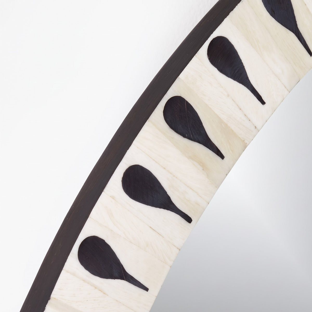 Close-up of a curved mirror frame with a light natural wood border, dark brown edge trim, and repeating black teardrop-shaped inlays, reflecting a light gray studio background.