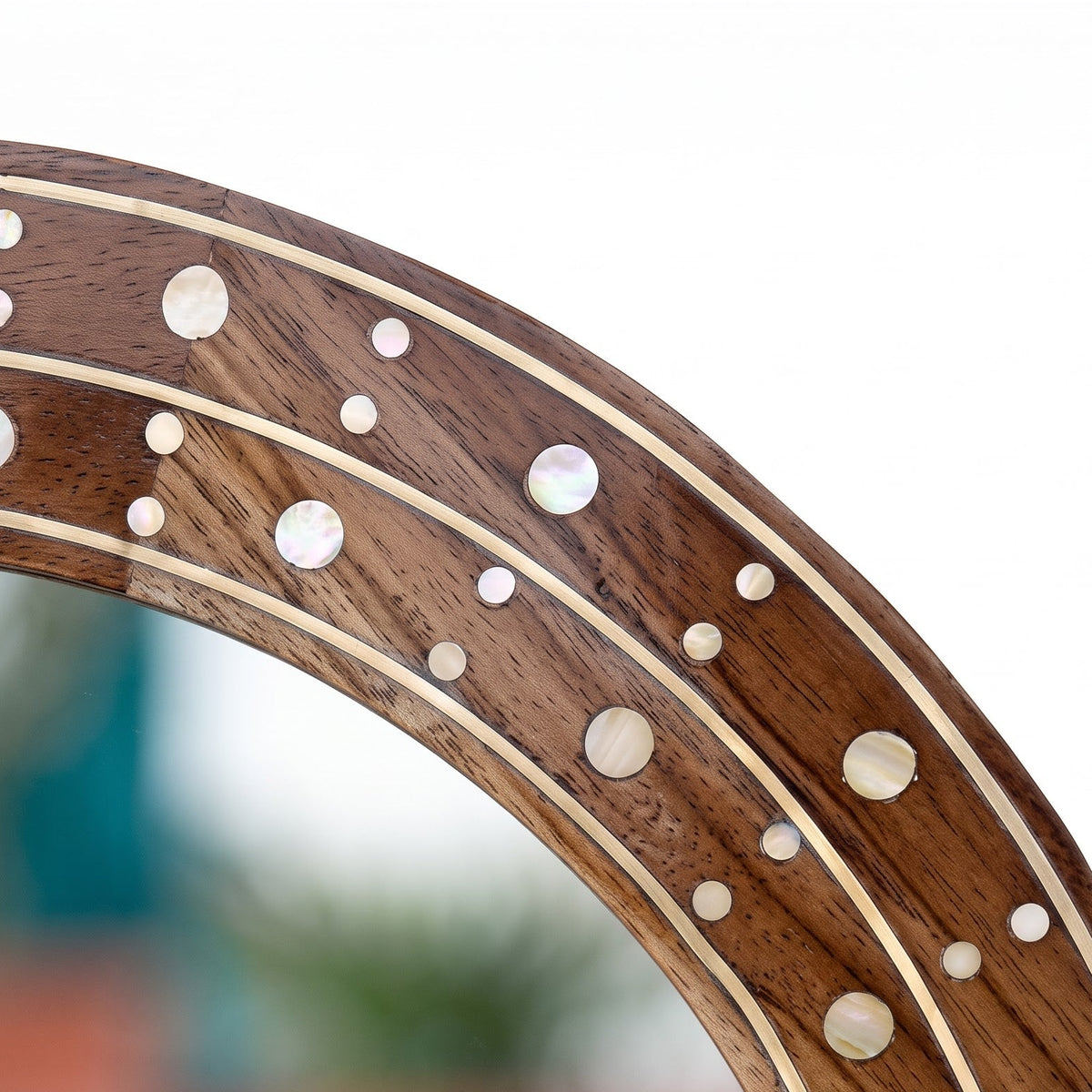 Close-up of a wooden mirror frame with inlaid mother-of-pearl circular accents and curved decorative stripes, shown against a blurred outdoor background.