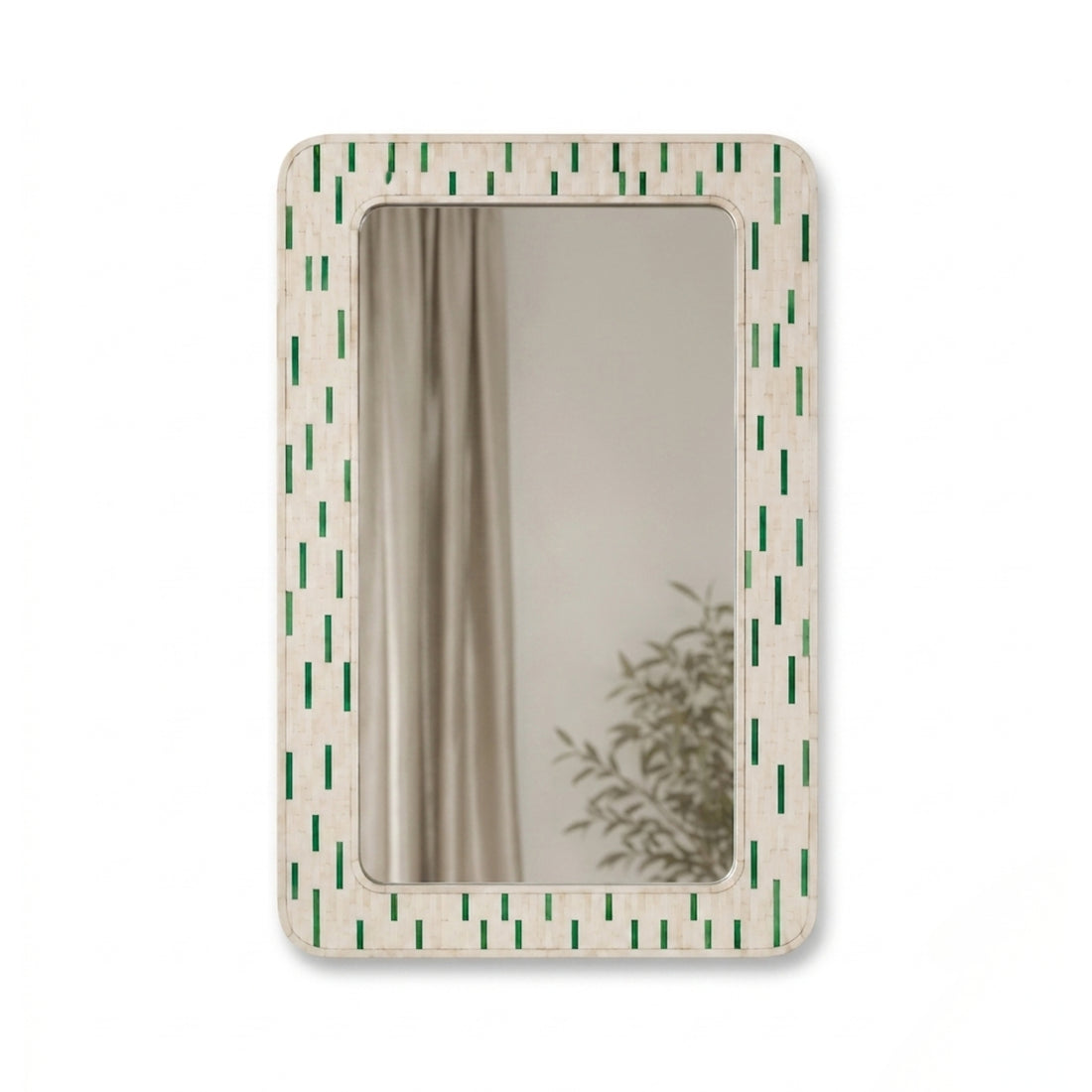 A rectangular wall mirror with a cream-colored frame featuring a vertical pattern of green dashes, styled on a wooden shelf next to a lit table lamp and a small vase of dried flowers.