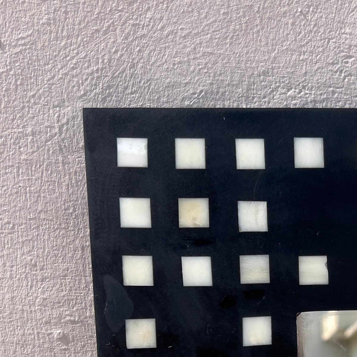 A close-up of a black rectangular frame with a filmstrip-style border of small white square inlays, leaning against a light gray textured wall. The mirror surface reflects an outdoor concrete courtyard and building wall.