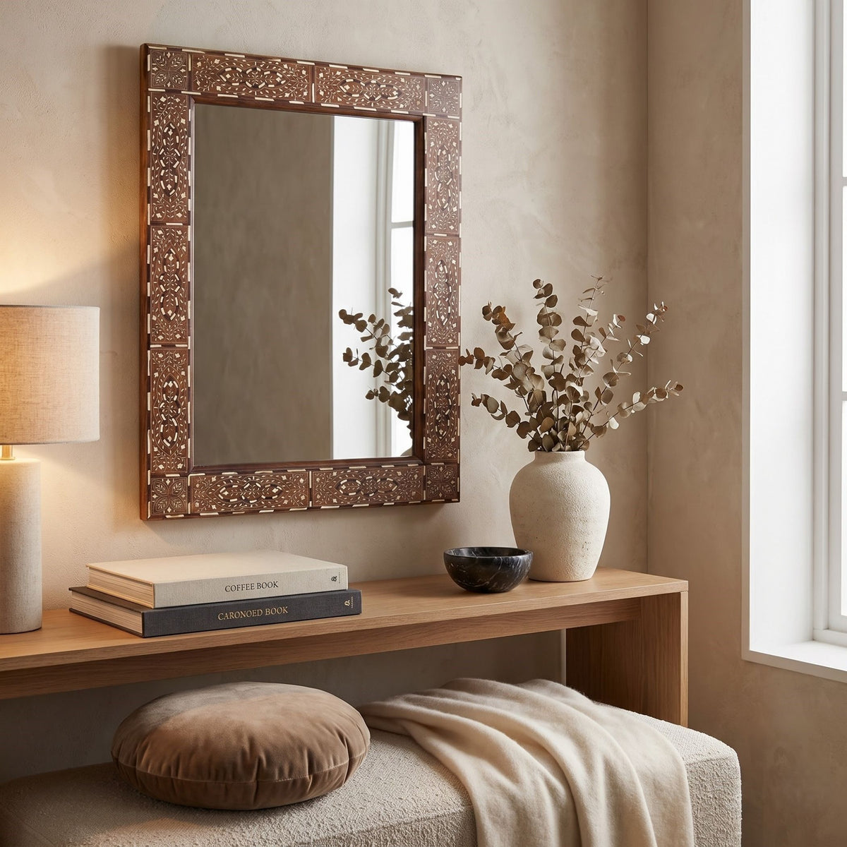 An ornate rectangular wall mirror with a carved brown wooden frame hangs above a light wood console table in a styled interior. The scene includes a lamp, books, a ceramic vase with dried branches, a small bowl, a cushion, and a throw blanket.