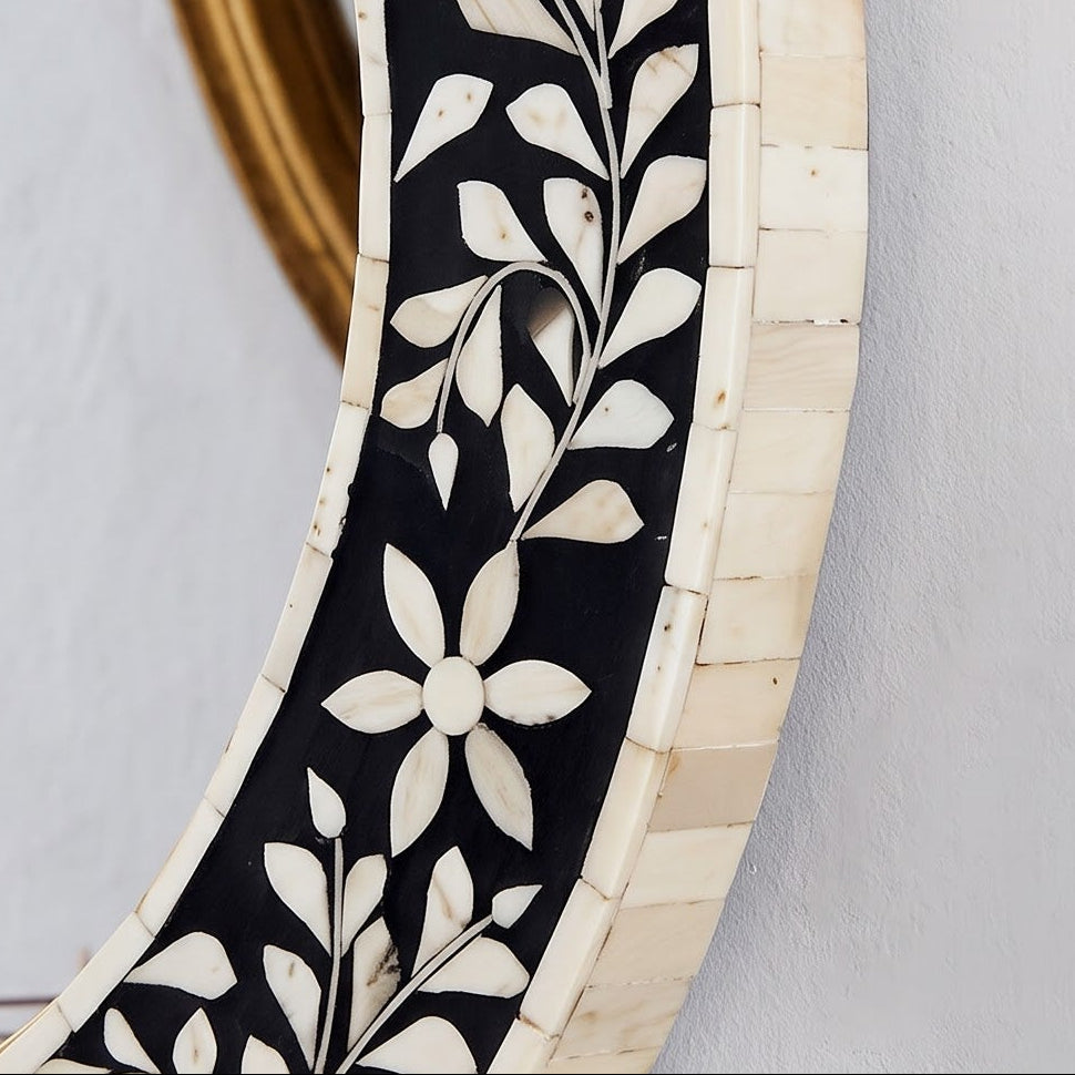 An oval decorative wall mirror with a black inlay featuring a cream floral and leaf pattern, a light natural wood frame, and a gold-toned inner rim. The mirror is mounted on a light gray textured wall, with its reflection showing a glass vase with a dried flower.