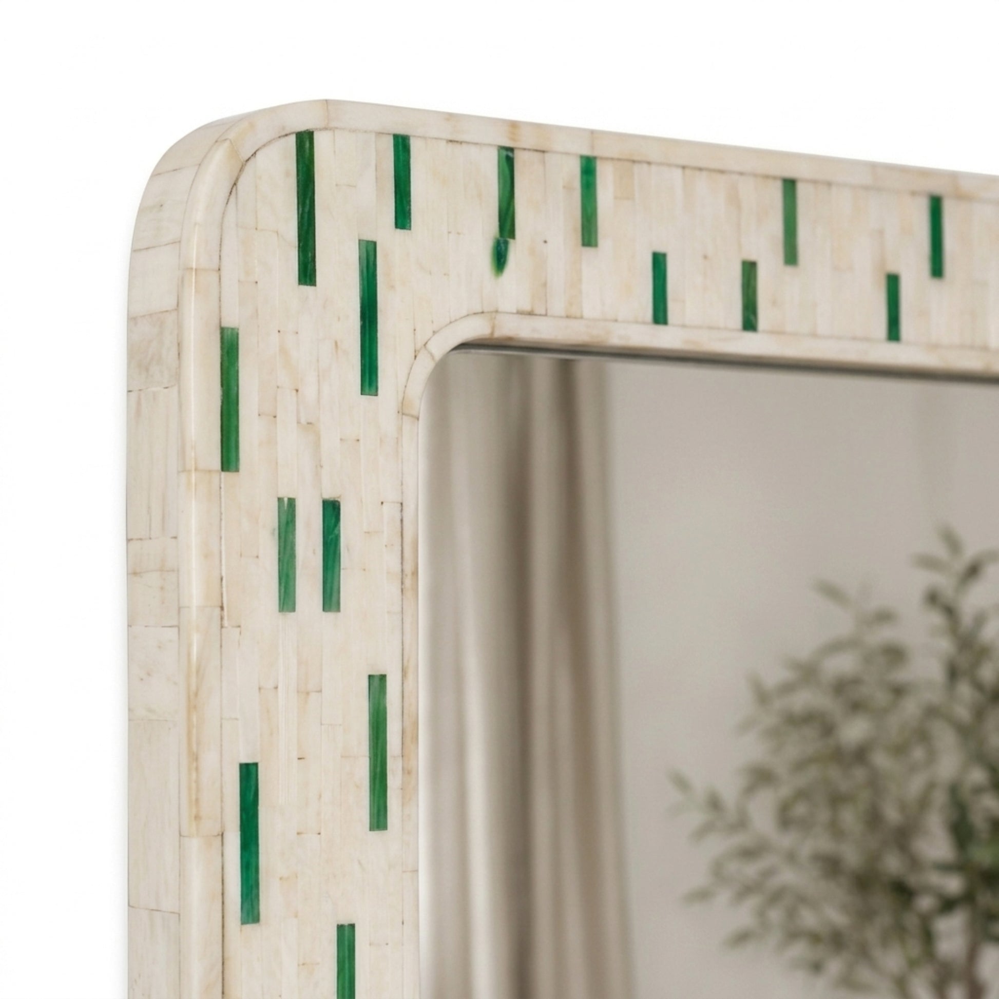 A close-up of a rectangular wall mirror with a light beige inlay frame decorated with vertical green mosaic accents, reflecting a softly blurred interior scene.