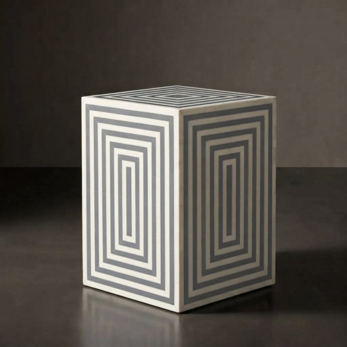 A square side table or stool with a geometric pattern of concentric rectangles in gray and off-white, set against a dark gray background.