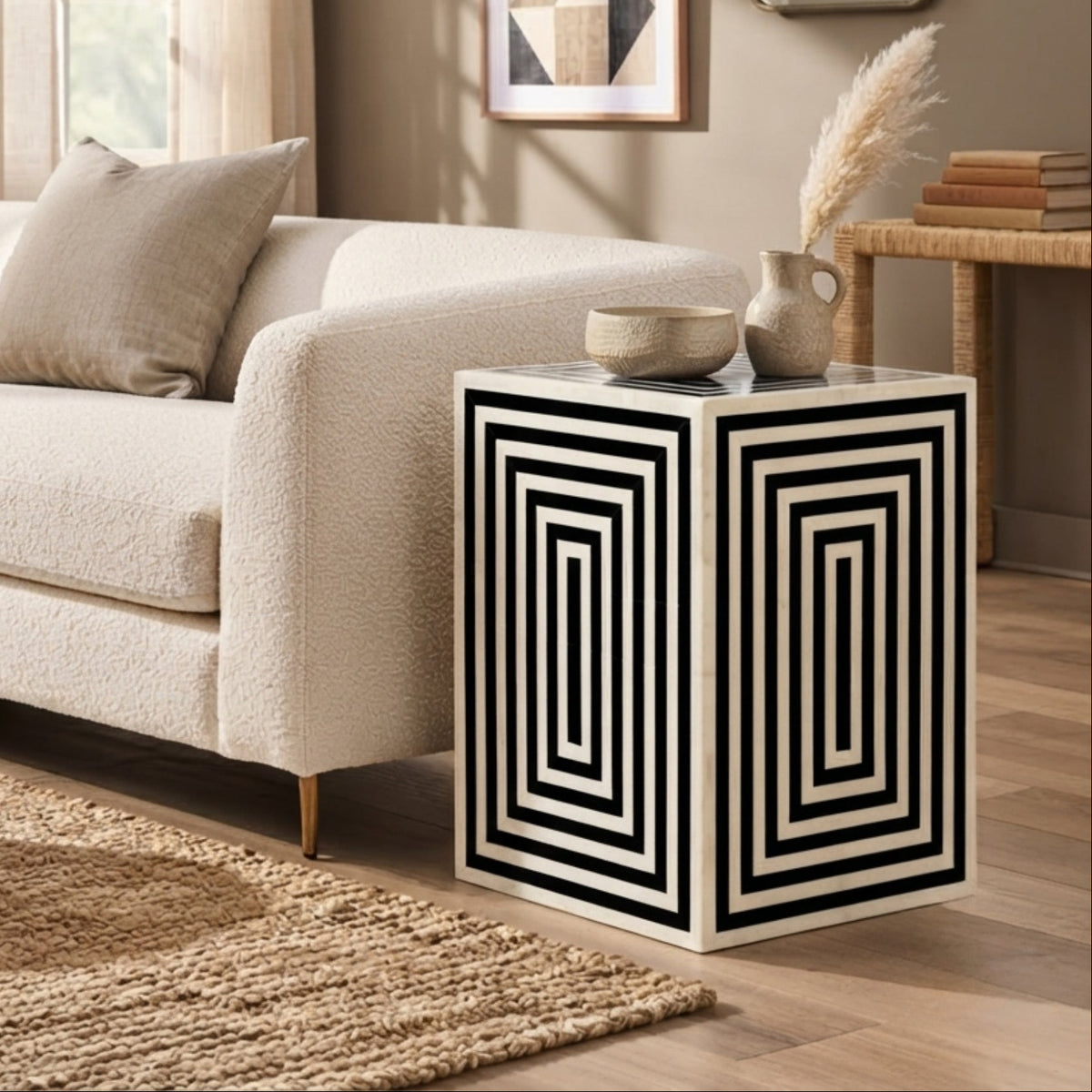 A modern side table with a bold black and white geometric concentric square pattern on its cube-shaped side, styled in a contemporary living room next to a cream bouclé sofa.