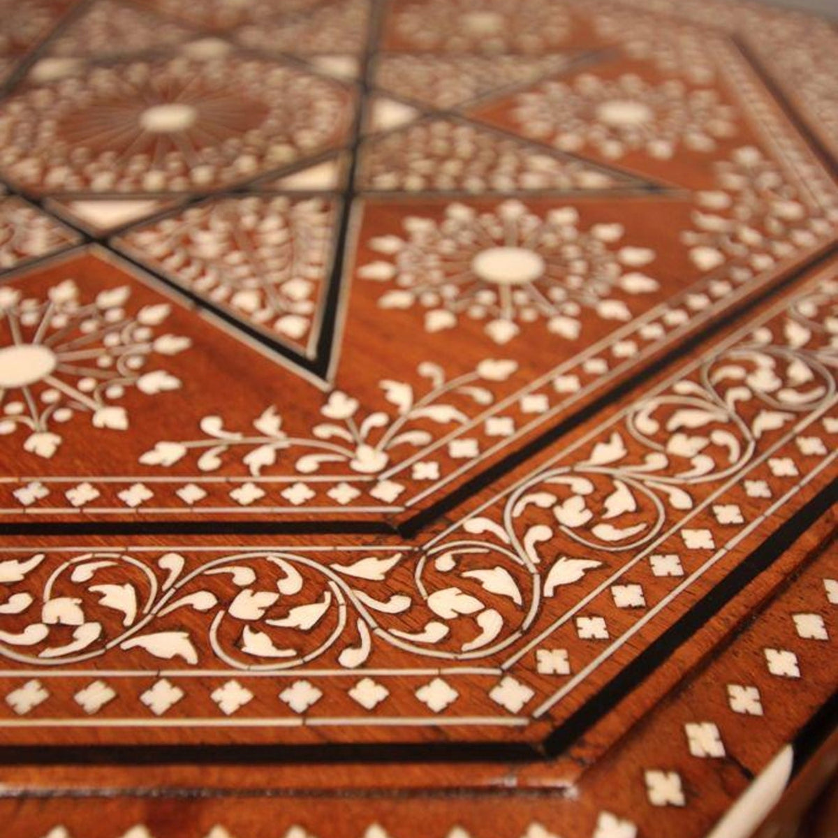 Close-up of an ornate geometric and floral inlay pattern on a reddish-brown wooden surface, with cream-colored decorative details forming stars, circles, and vine motifs.