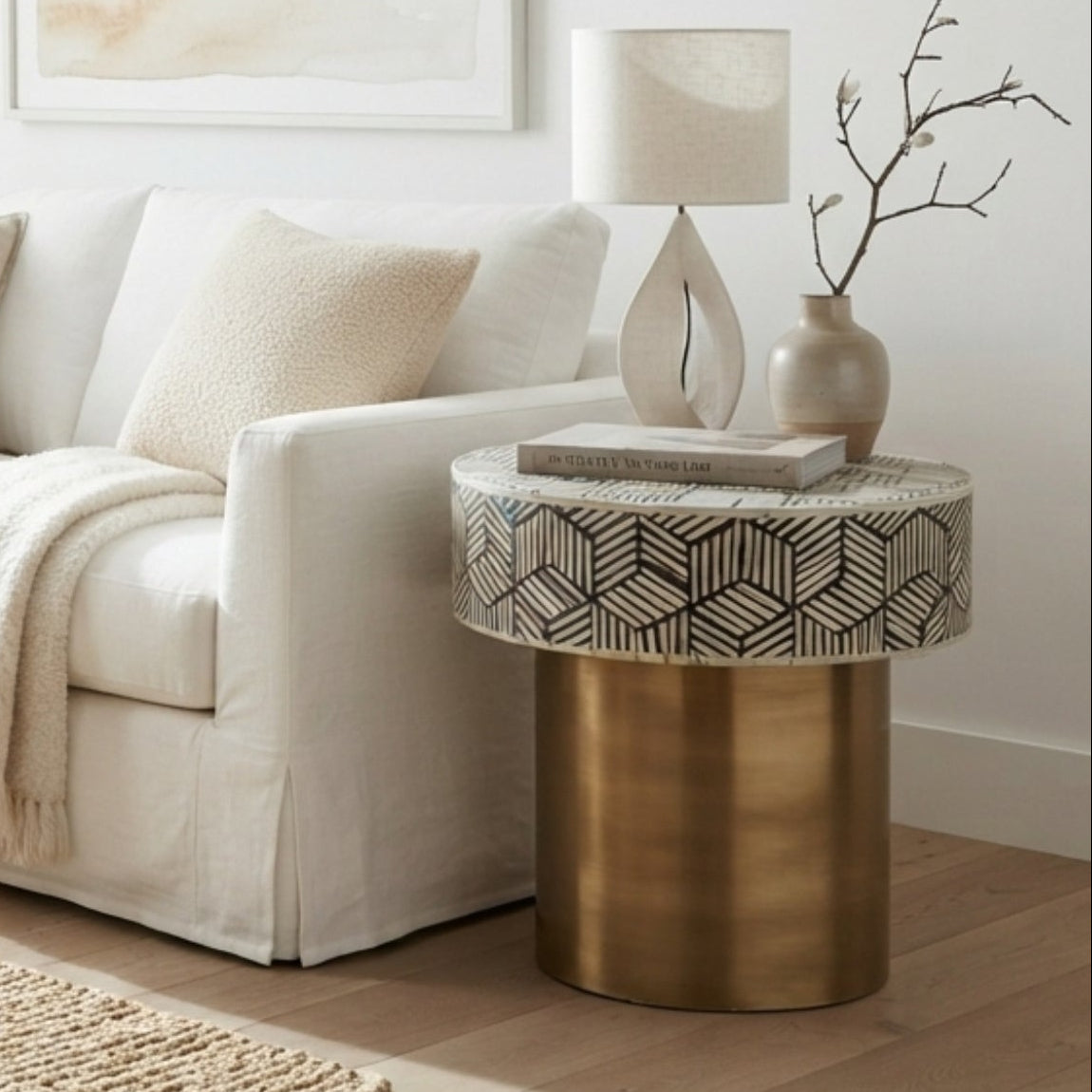 A styled living room scene featuring a cream slipcovered sofa with neutral throw pillows, a woven rattan armchair, a round gold side table with a patterned top, and a large natural jute rug on a light wood floor.