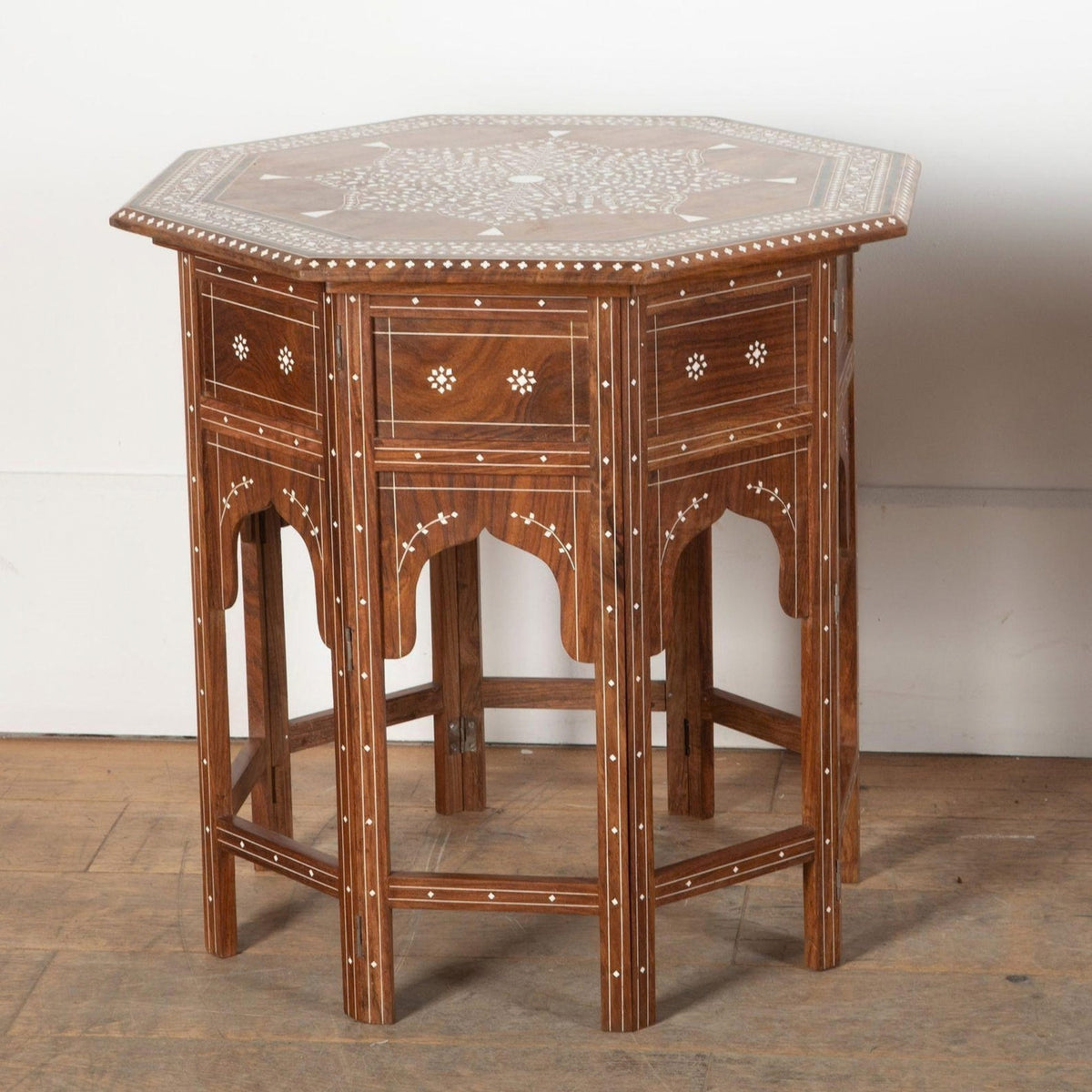 An octagonal wooden side table with intricate inlay detailing, featuring three drawers on the top, decorative arches on the front, and a geometric patterned top surface, set against a plain white wall and wooden floor.