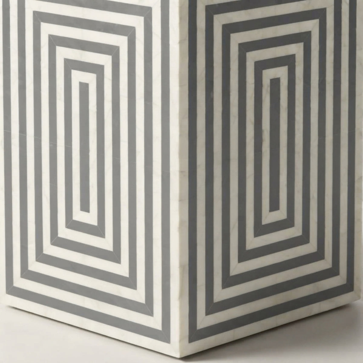 A close-up of a geometric side table or box with a white base and alternating dark gray concentric rectangular inlay pattern.