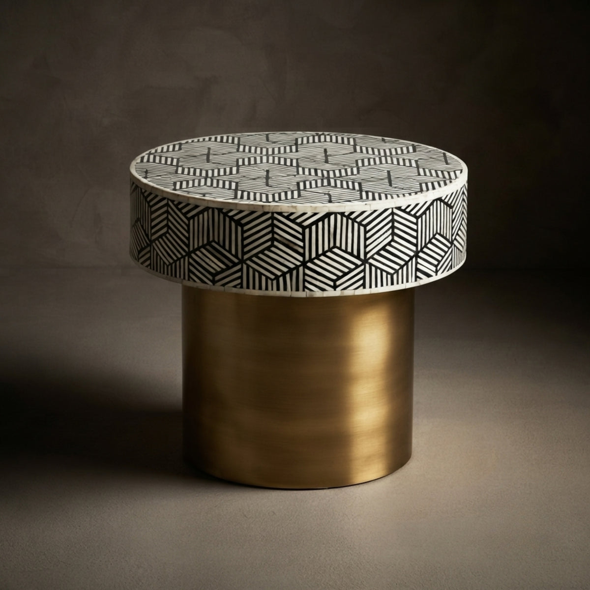A modern cylindrical side table with a brushed gold metal base and a round top featuring a black and white geometric bone-inlay pattern, set against a dark textured background.
