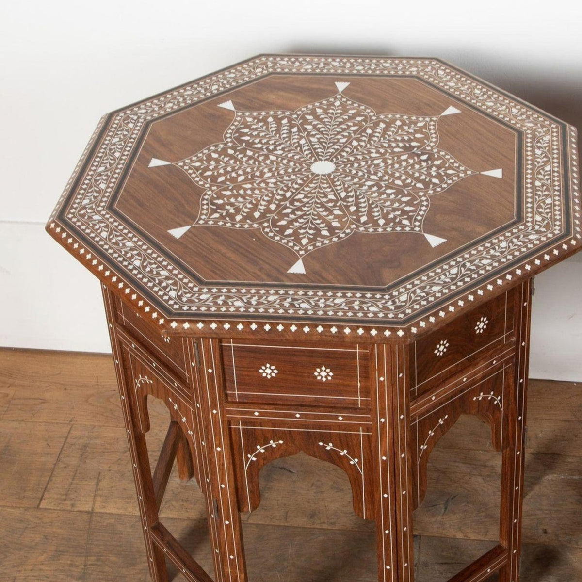 An octagonal wooden side table with intricate white inlay detailing on the tabletop and drawer fronts, set against a plain white wall and wooden floor.