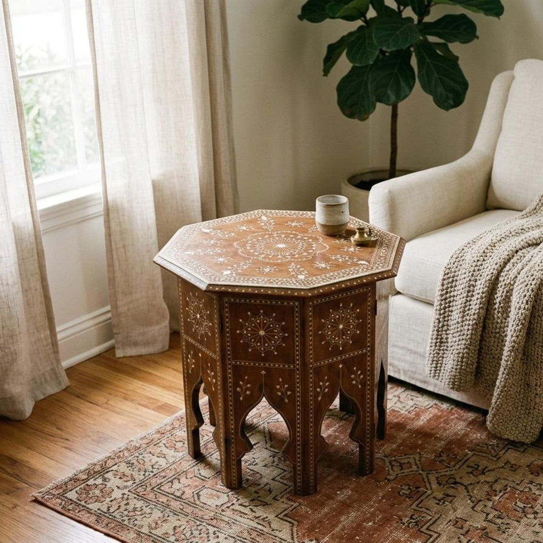 An octagonal wooden end table with intricate bohemian-style inlay patterns, styled in a cozy living room next to a white armchair and a potted fiddle-leaf fig plant.