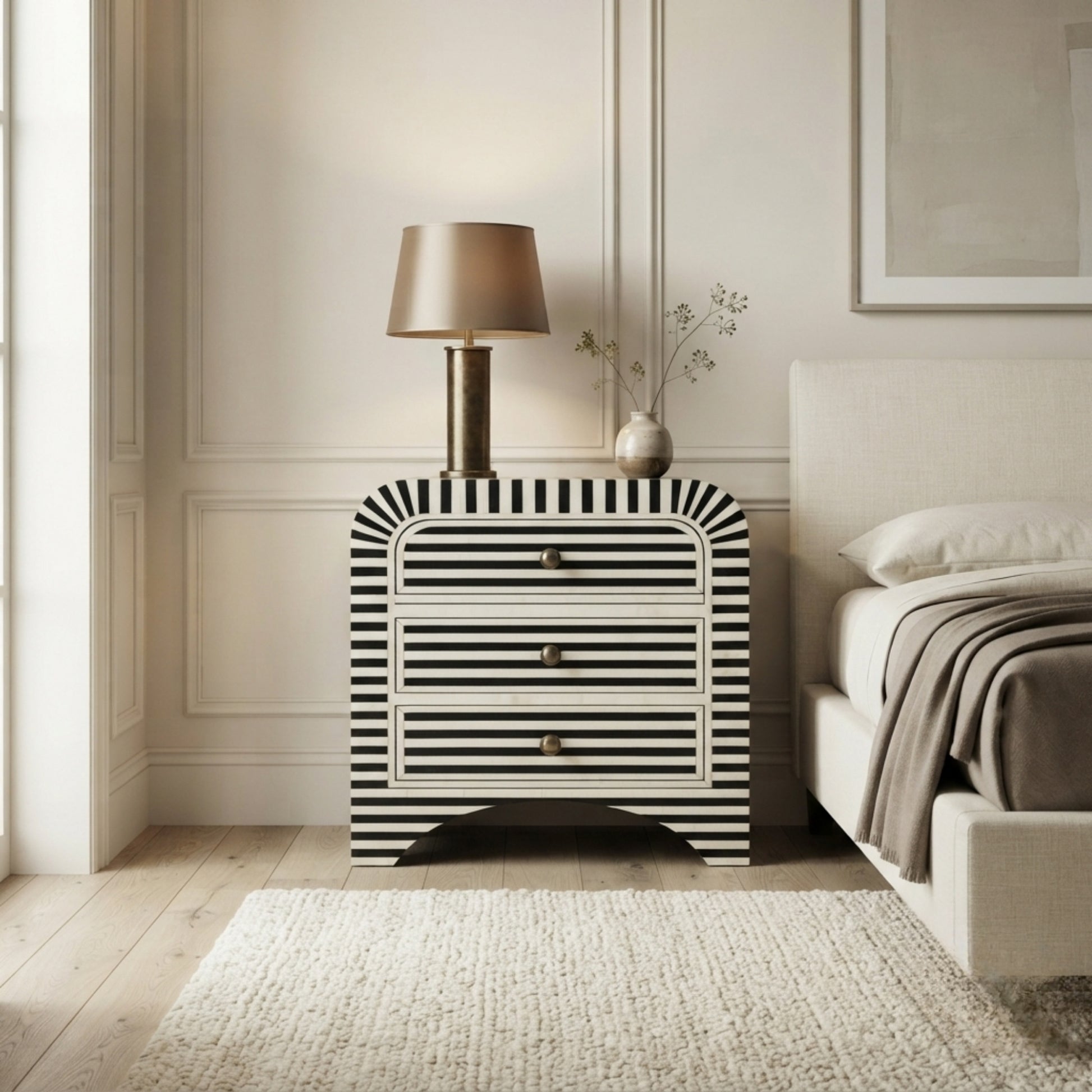 A modern three-drawer nightstand with a bold black and white striped pattern and curved top, styled in a bedroom next to a bed with a lamp on top.