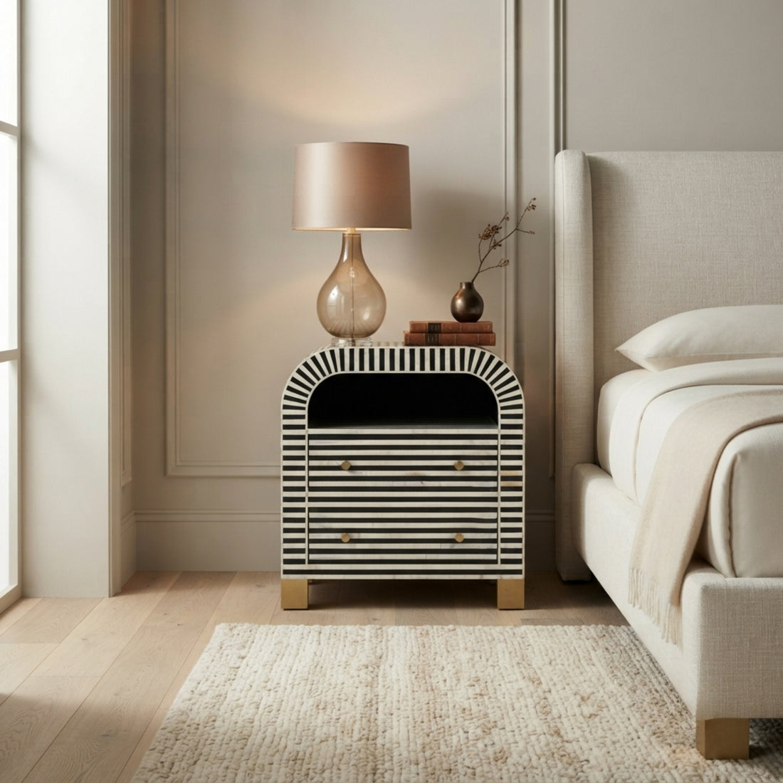 A modern bedside table with a black and white striped bone-in-laminate finish, styled in a bedroom next to a bed with a lit table lamp on top.