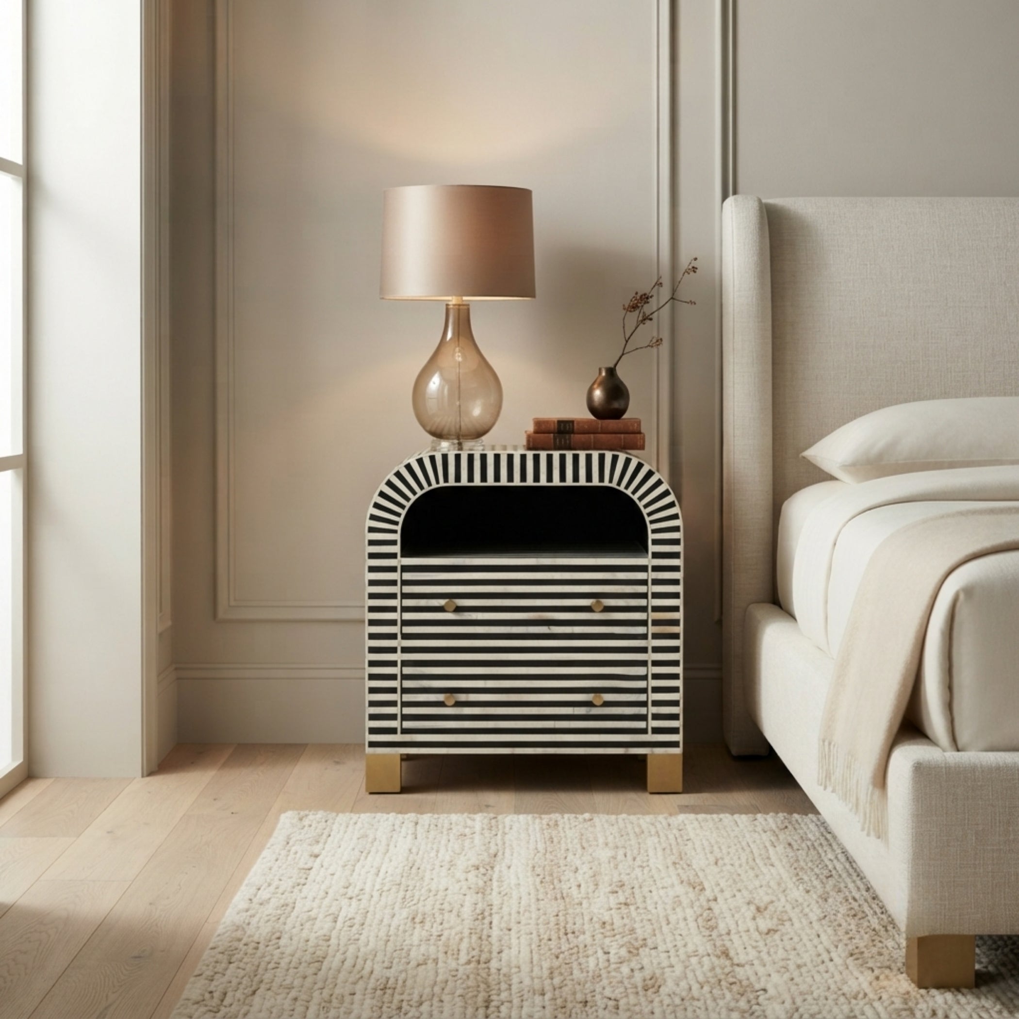 A modern bedside table with a black and white striped bone-in-laminate finish, styled in a bedroom next to a bed with a lit table lamp on top.