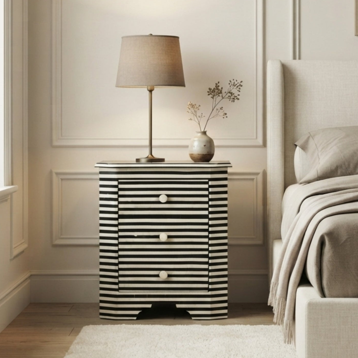 A modern bedside table with black and white horizontal stripes, styled in a bedroom next to a bed with neutral beige bedding and a table lamp.