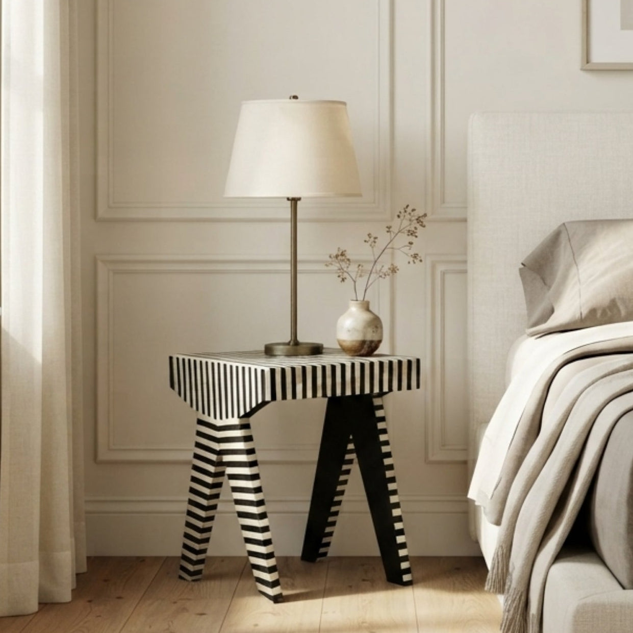 A modern bedroom scene featuring a bed with neutral beige and gray bedding, a black and white striped side table with a lamp, light wood floors, and white paneled walls.