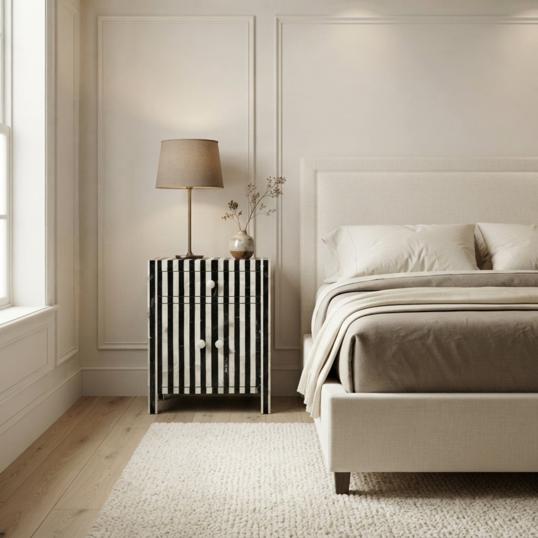 A modern bedroom scene featuring a black and white vertically striped nightstand with a table lamp and vase on top, positioned next to a bed with neutral beige bedding and a textured rug.