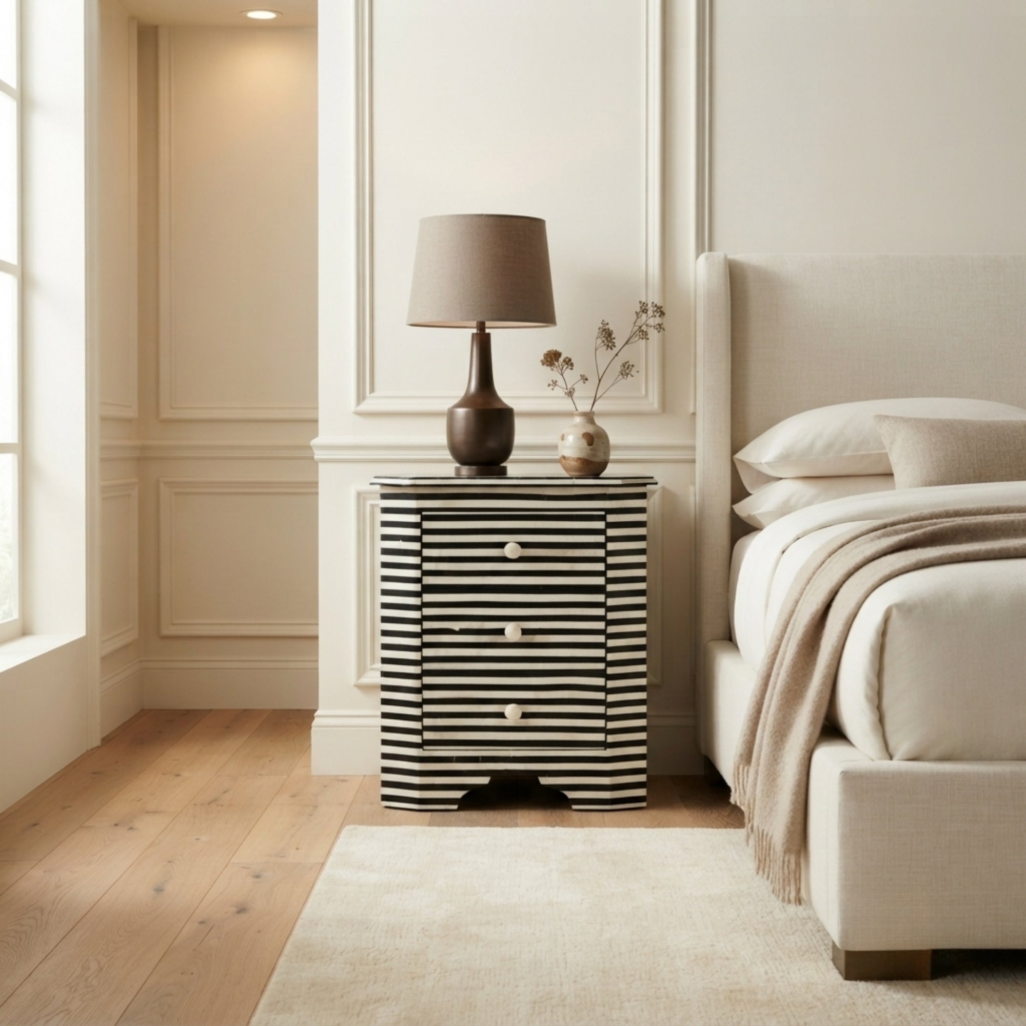 A modern bedside table with black and white horizontal stripes, styled in a bright, neutral bedroom next to a bed with beige bedding.