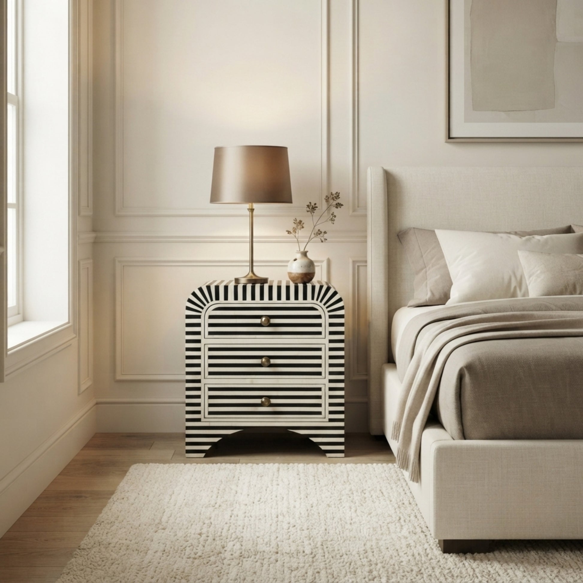 A modern bedroom scene featuring a black and white striped nightstand with a table lamp and small vase on top, positioned next to a bed with neutral beige bedding.