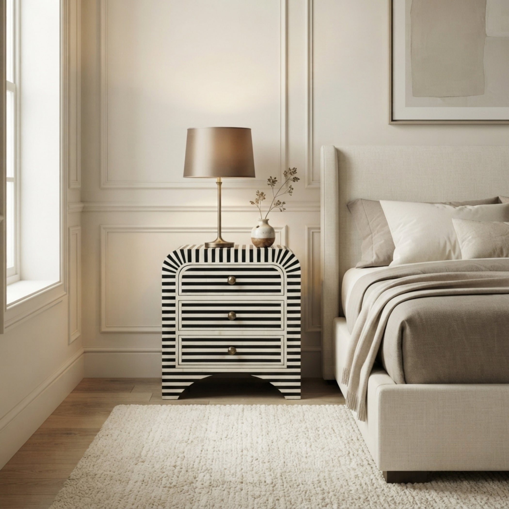 A modern bedroom scene featuring a black and white striped nightstand with a table lamp and small vase on top, positioned next to a bed with neutral beige bedding.