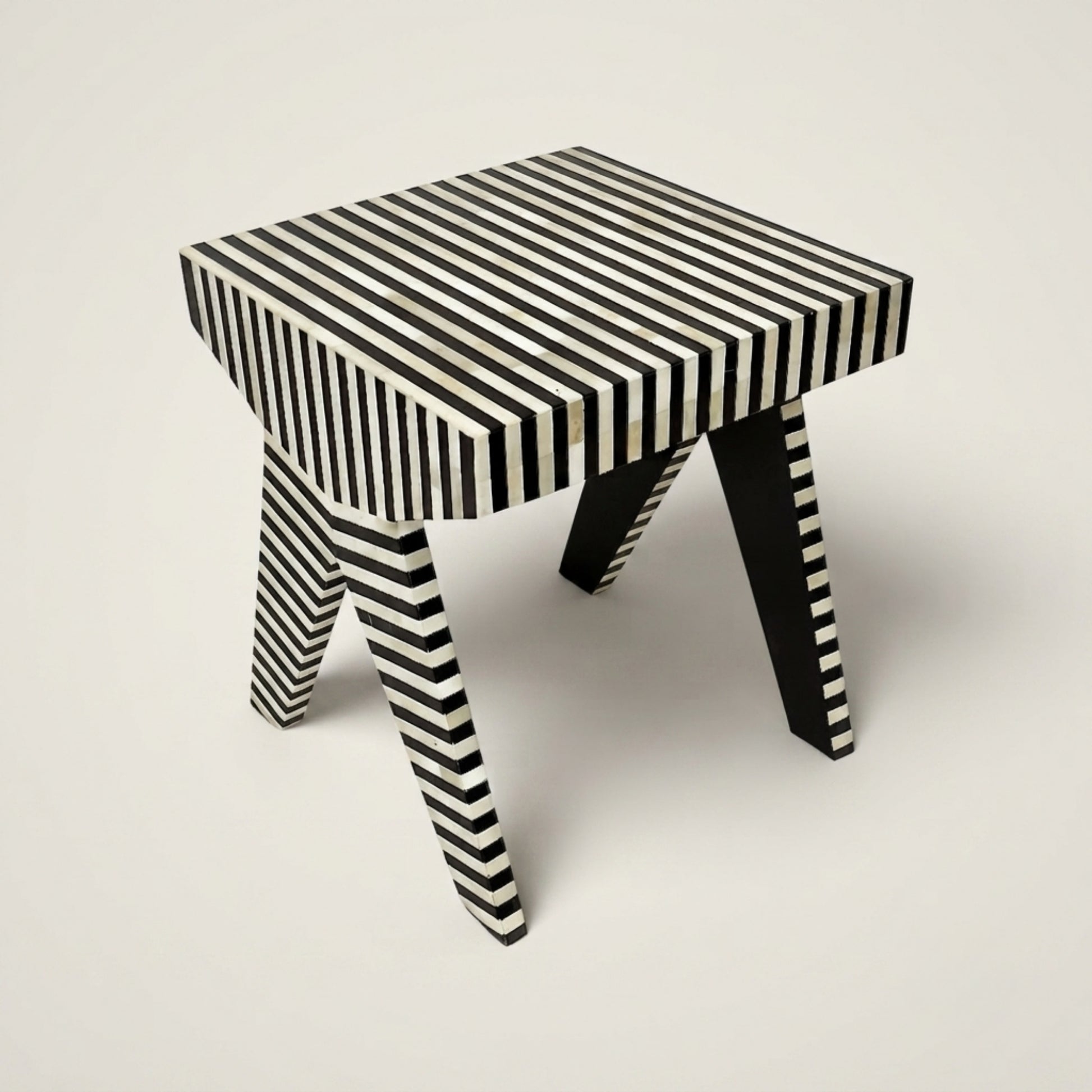A small stool with a black and white striped pattern, featuring a square top and angled legs, photographed on a neutral light gray background.