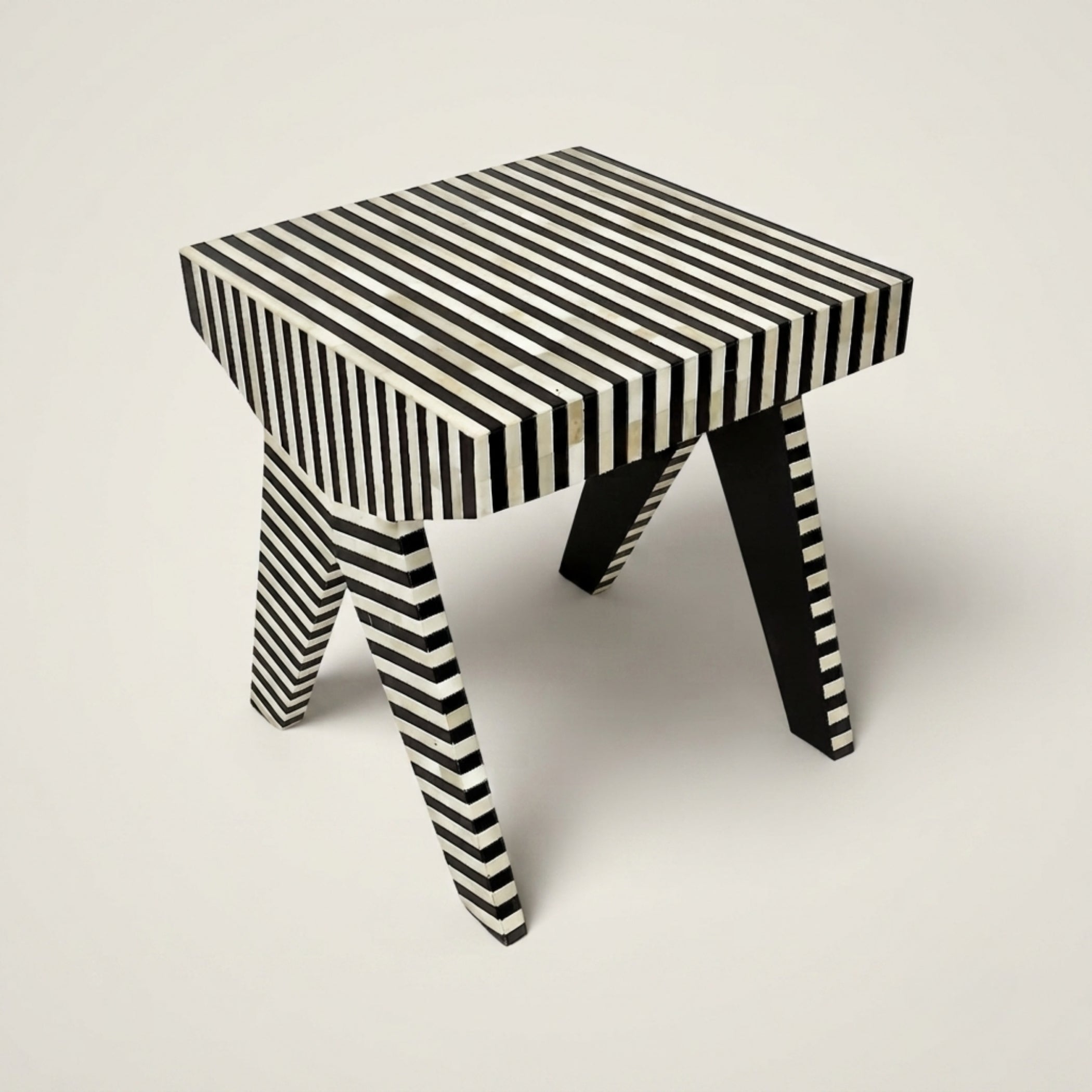 A small stool with a black and white striped pattern, featuring a square top and angled legs, photographed on a neutral light gray background.
