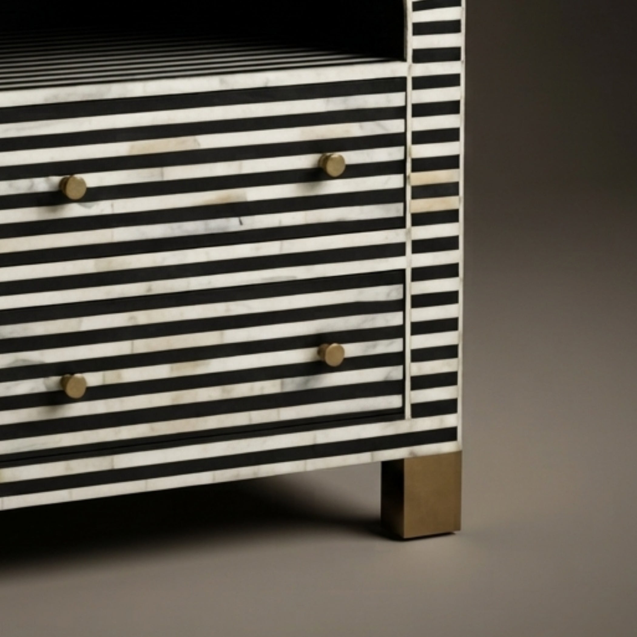 Close-up of a striped furniture piece with black and white horizontal inlay, small round brass knobs, and a brass leg on a neutral studio background.