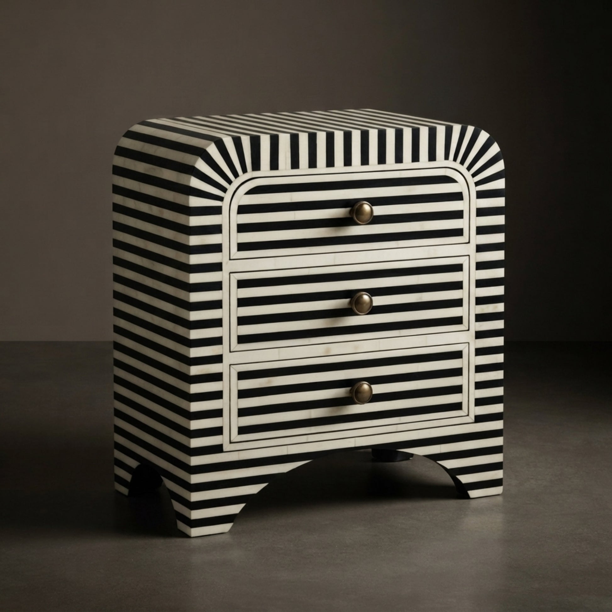 A three-drawer nightstand with a bold black and white striped pattern and curved edges, featuring three drawers with round gold-toned knobs, set against a dark gray studio background.