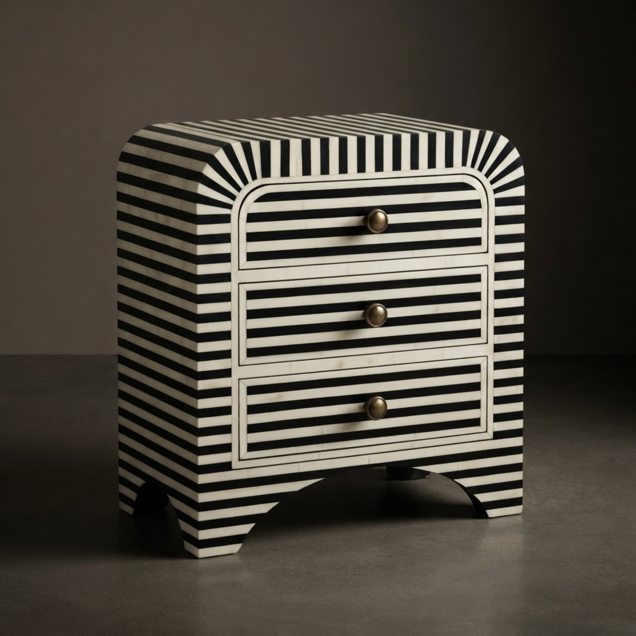 A three-drawer nightstand with a bold black and white striped pattern and curved edges, featuring three drawers with round gold-toned knobs, set against a dark gray studio background.