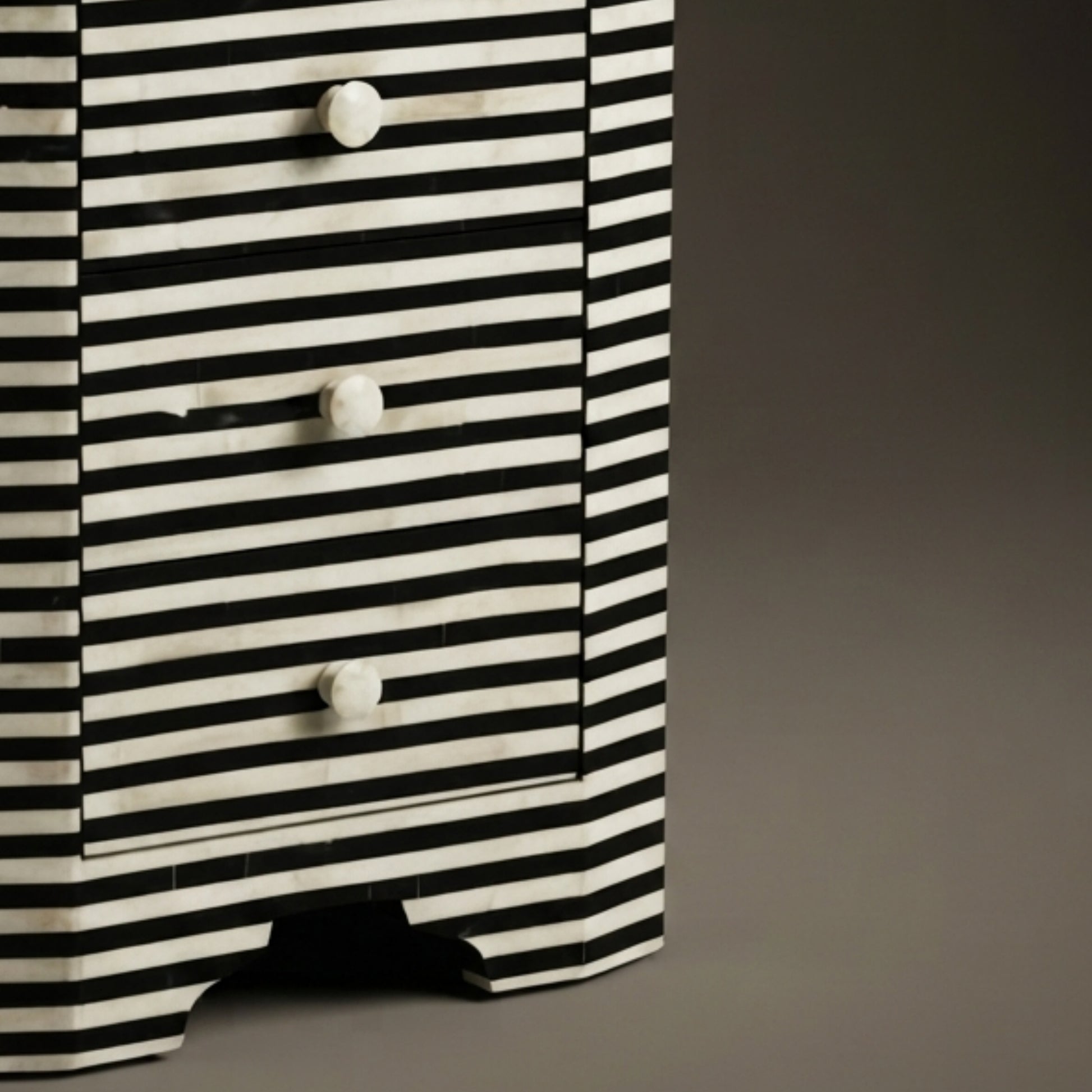 A close-up of a black and white striped wooden chest of drawers with three drawers and round white knobs, set against a dark gray background.