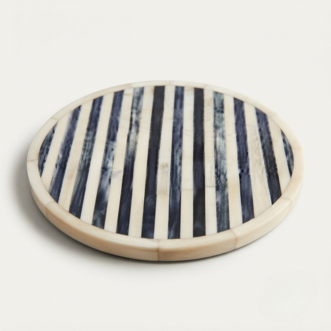 A small ceramic cup filled with amber tea sits on a round coaster with dark navy blue and cream vertical stripes, set on a textured off-white surface with soft natural shadows.