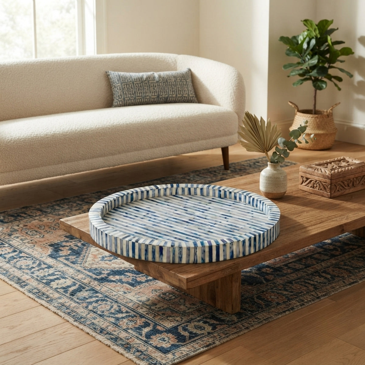 A round woven tray with blue and white striped cushions sits on a wooden coffee table in a bright, modern living room with a cream boucle sofa and patterned rug.