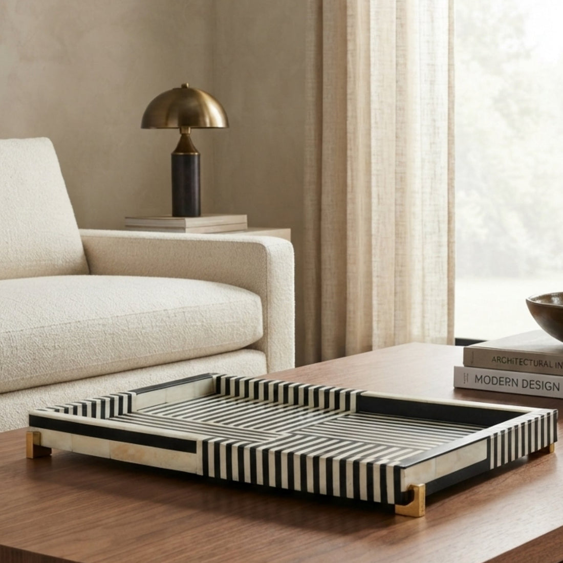 A rectangular serving tray with black and white striped bone-look sides and gold-toned feet, styled on a wooden coffee table in a modern living room setting.