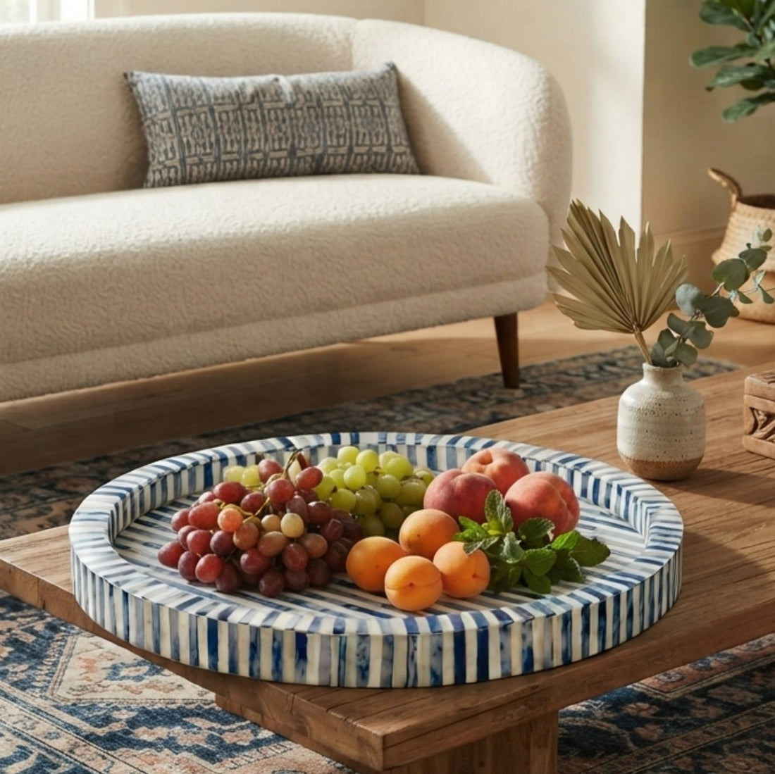 A round woven tray with blue and white stripes, filled with fruit, placed on a wooden coffee table in a bright, modern living room.