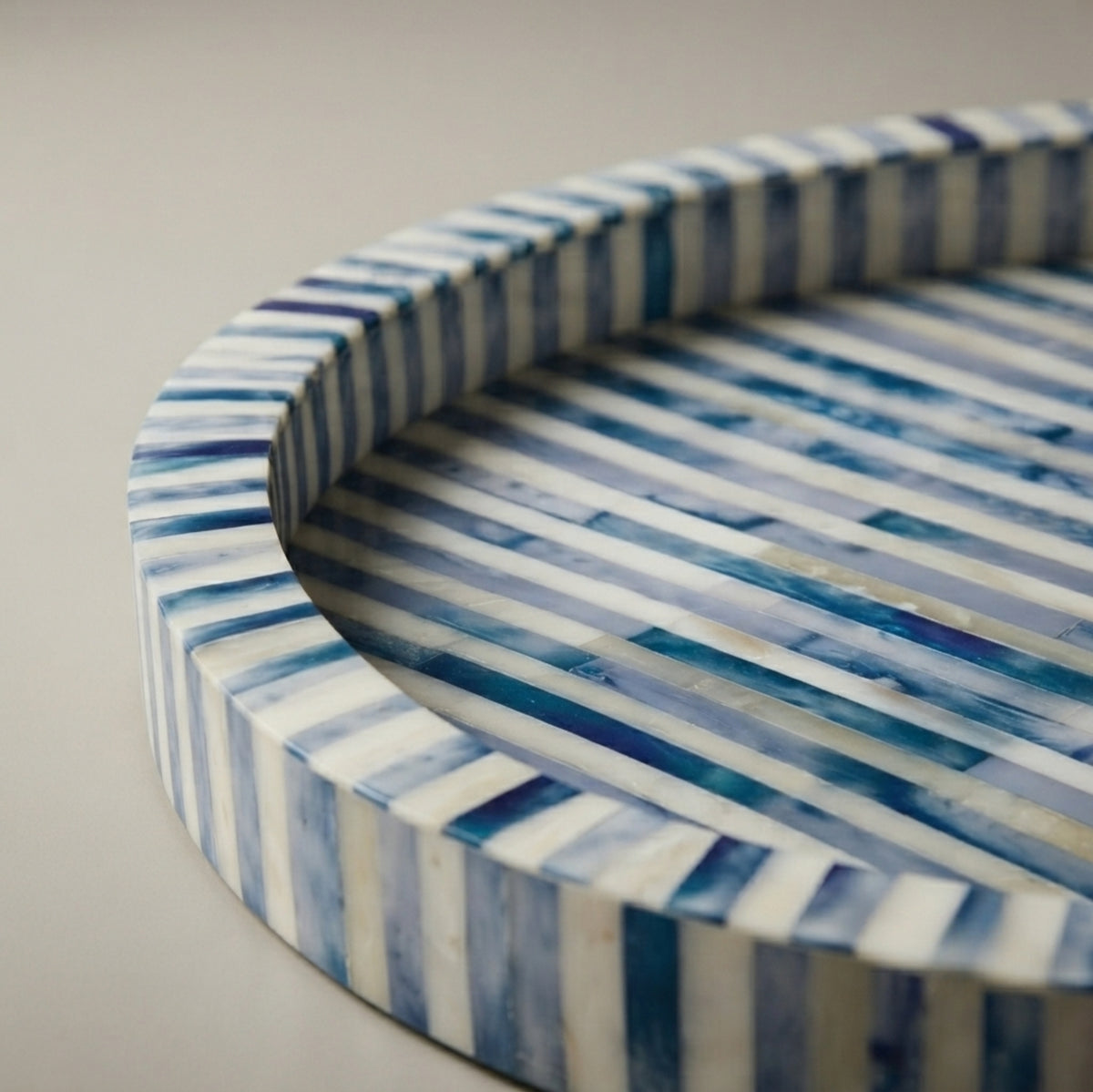 A round decorative tray with a striped pattern of blue and white bone-insect inlay, shown in a close-up view on a neutral light gray background.