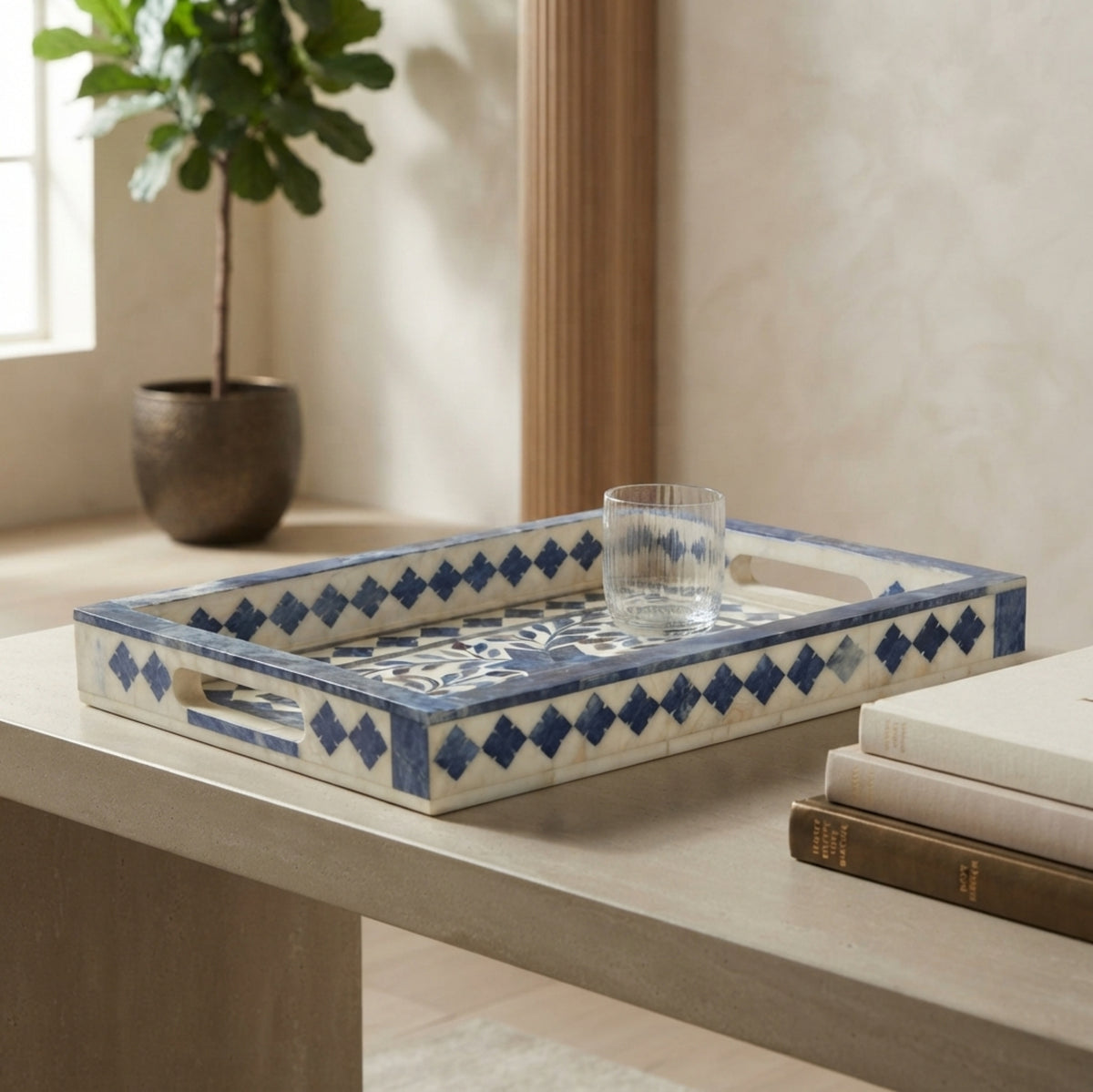 A rectangular decorative tray with blue and white geometric patterns sits on a light wood table in a styled interior setting, with a glass cup placed on top and books nearby.