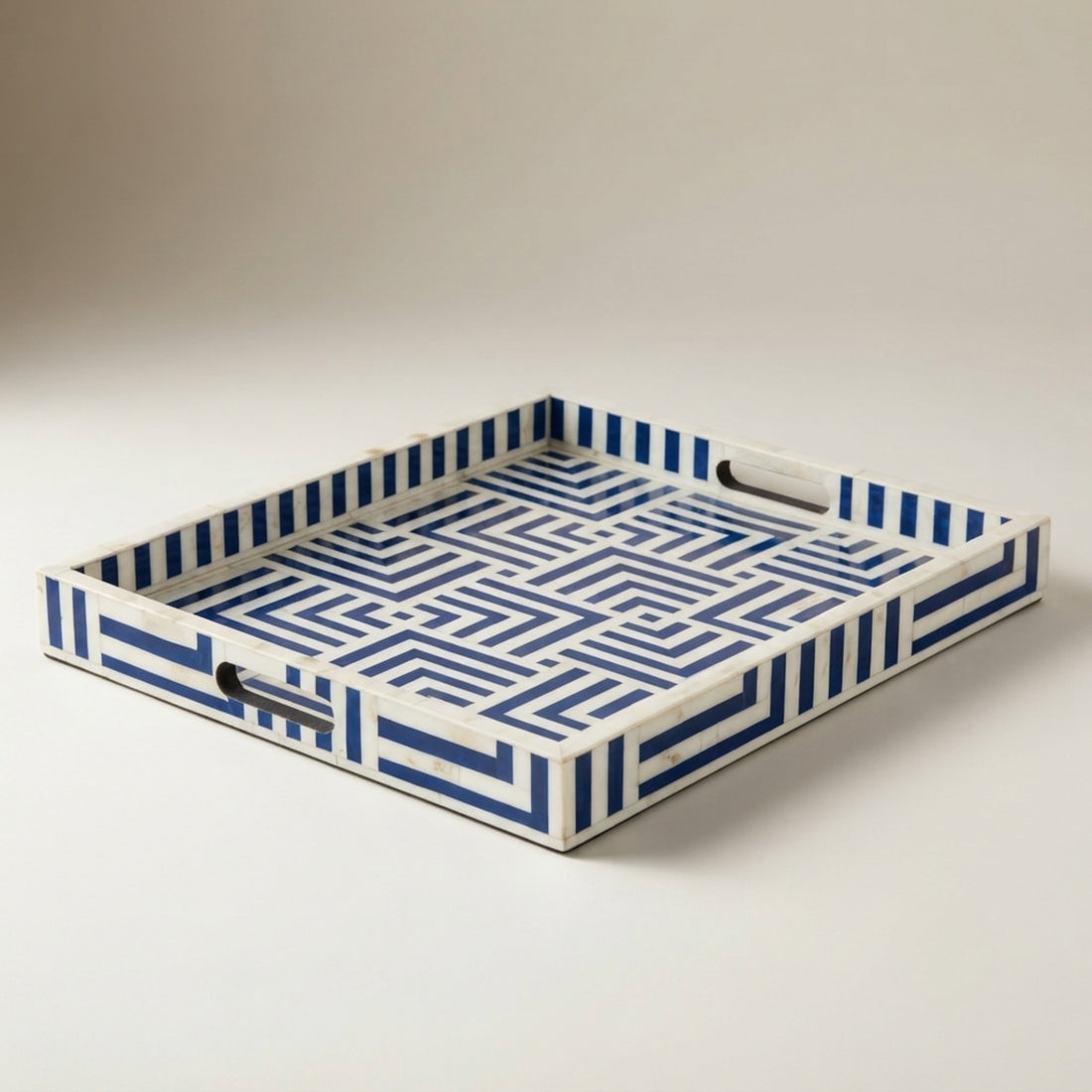 A rectangular decorative tray with a white base and bold blue geometric border pattern, styled on a dark table in a bright room. The tray holds gold candlesticks, a clear textured bottle, a small white box, and gold bells, with a chair and vase visible in the background.