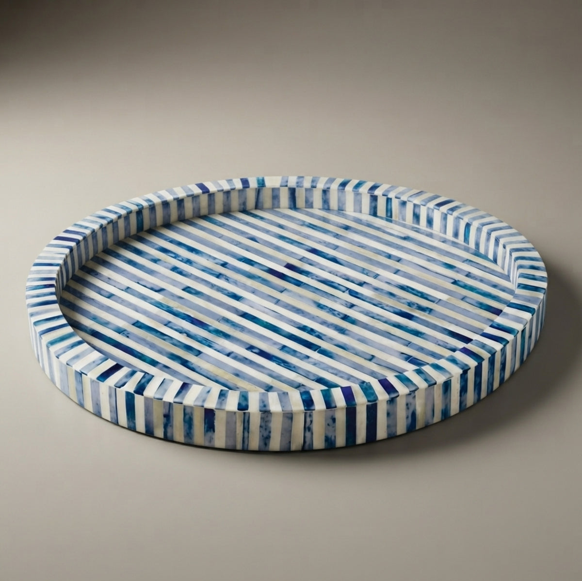 Round decorative tray with diagonal blue and white striped inlay, photographed on a neutral warm gray background.