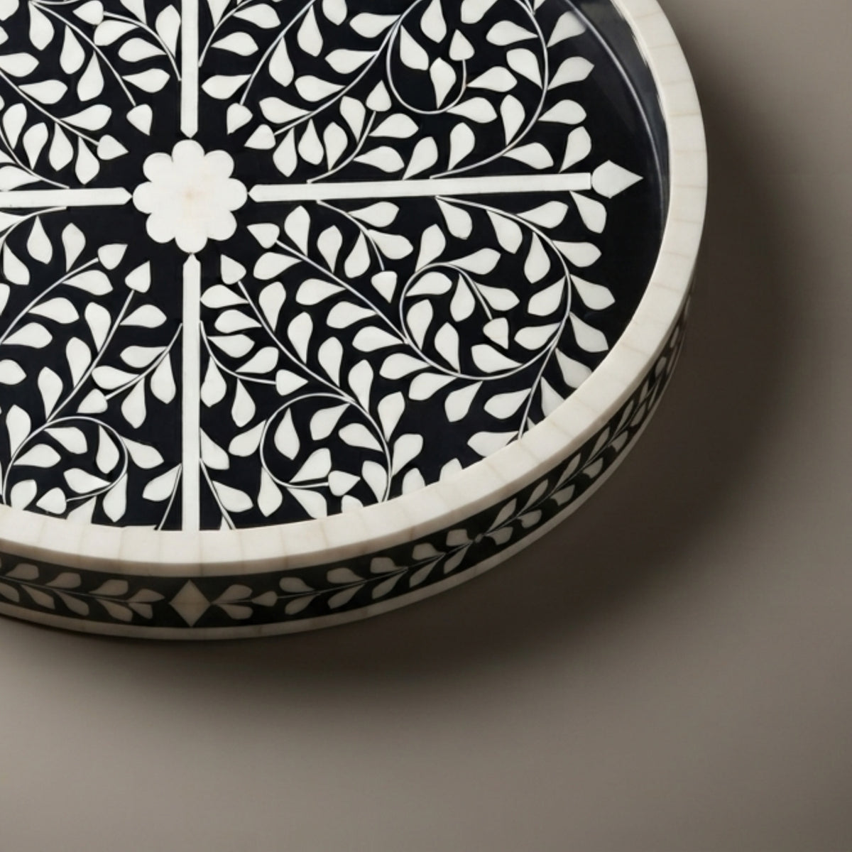 A close-up of a round decorative tray or dish with a black and white floral inlay pattern, photographed on a neutral taupe background.