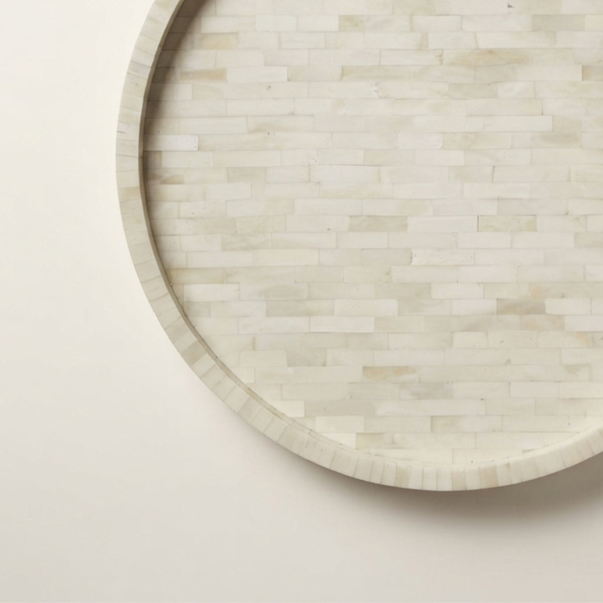 A close-up of a round tray made from small rectangular bone or shell pieces in a light ivory to beige tone, photographed against a plain off-white background.
