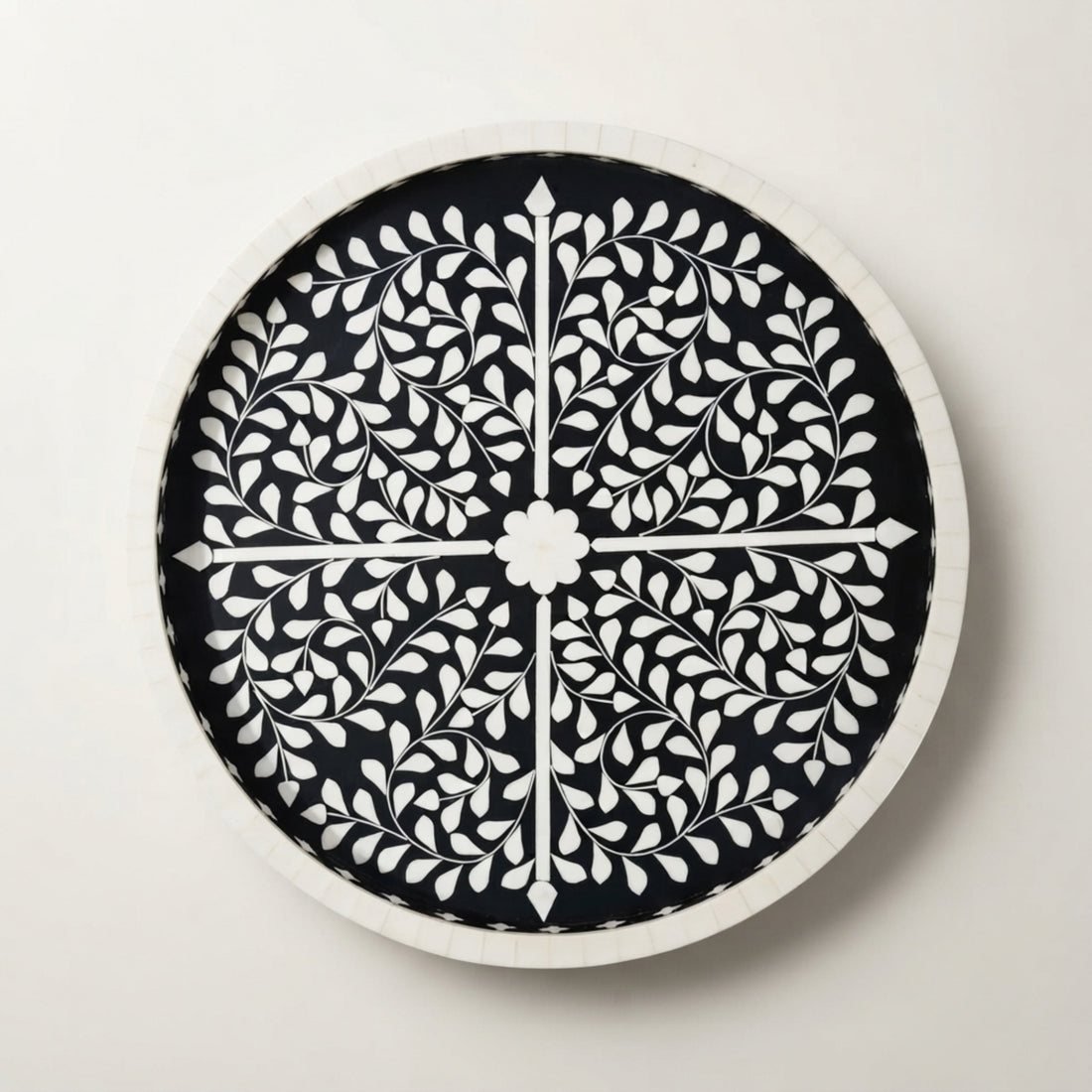 A round black and white patterned bone-inlay serving tray sits on a wooden coffee table in a beautifully styled living room with a white sofa, patterned throw pillows, a potted plant, and books.