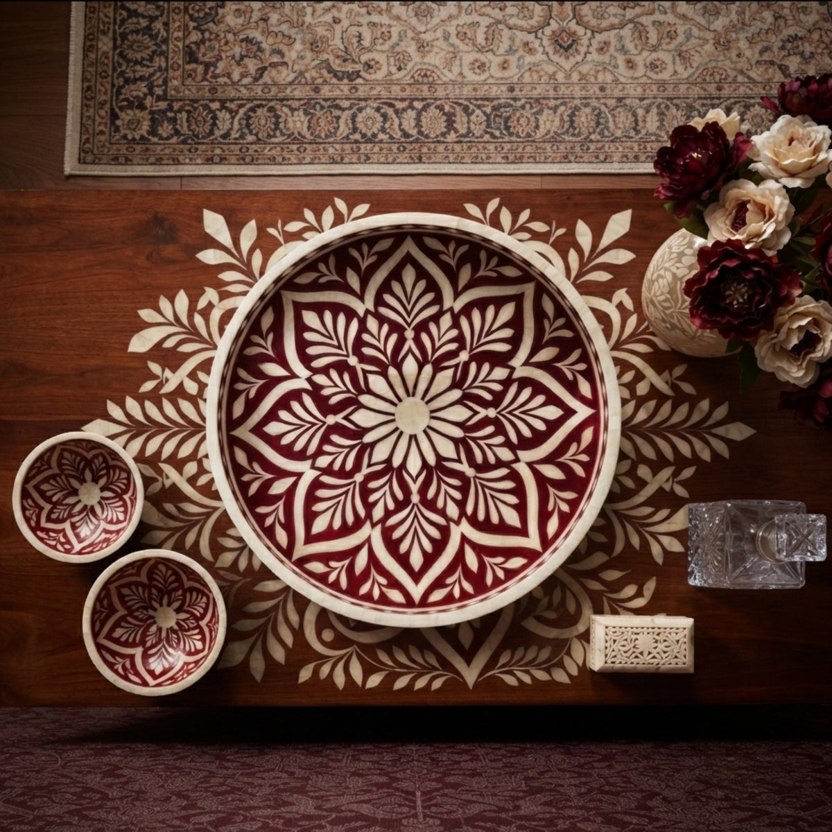 A set of hand-painted ceramic serving pieces, including a large round platter and two small bowls, featuring a detailed red and cream floral mandala pattern, arranged on a wooden table with decorative props.