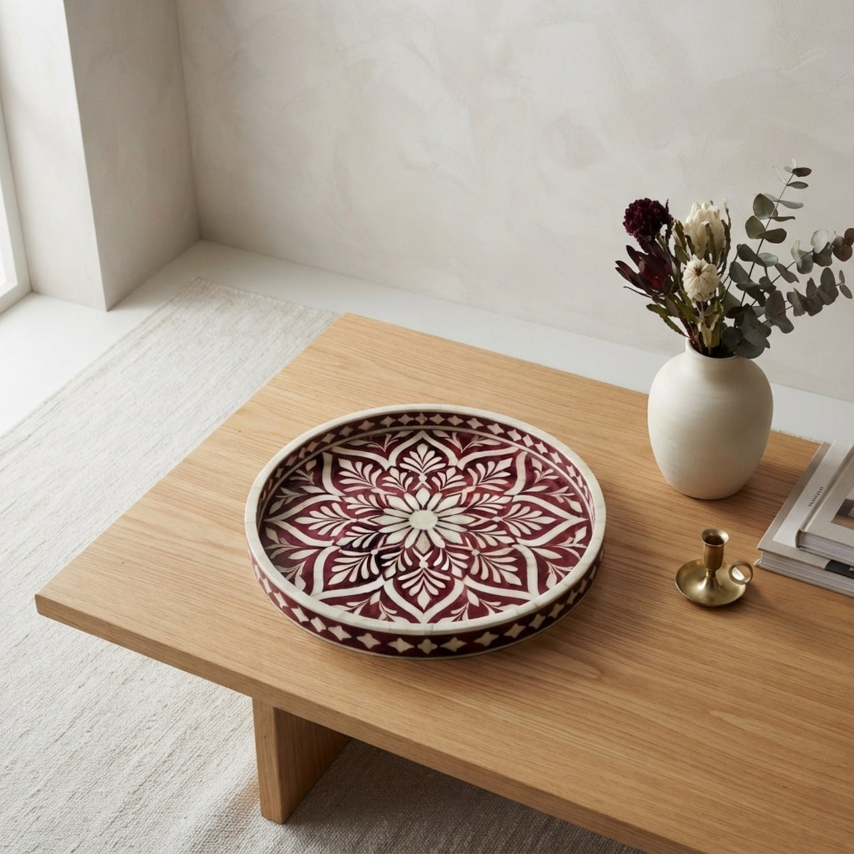 A round decorative tray with an intricate red and cream mandala pattern, styled on a light wood coffee table next to a white ceramic vase with dried flowers.