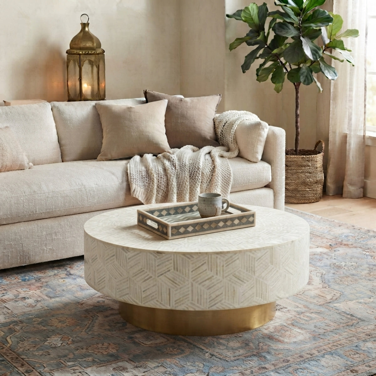 A round coffee table with a cream and gold geometric patterned top and a gold base, styled in a cozy living room next to a beige sofa.