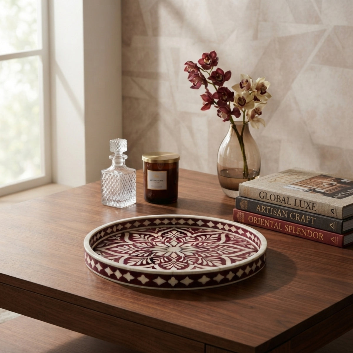 A round decorative tray with a red and cream mandala pattern sits on a wooden table in a styled room setting, alongside a glass decanter, a candle, and a vase with flowers.