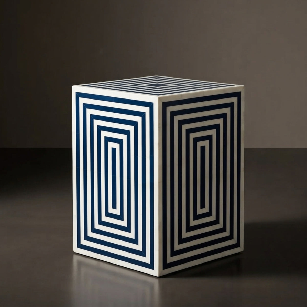 A cube-shaped side table or stool with a white base and dark navy blue geometric concentric line patterns on two visible sides, photographed in a studio setting against a dark gray background.