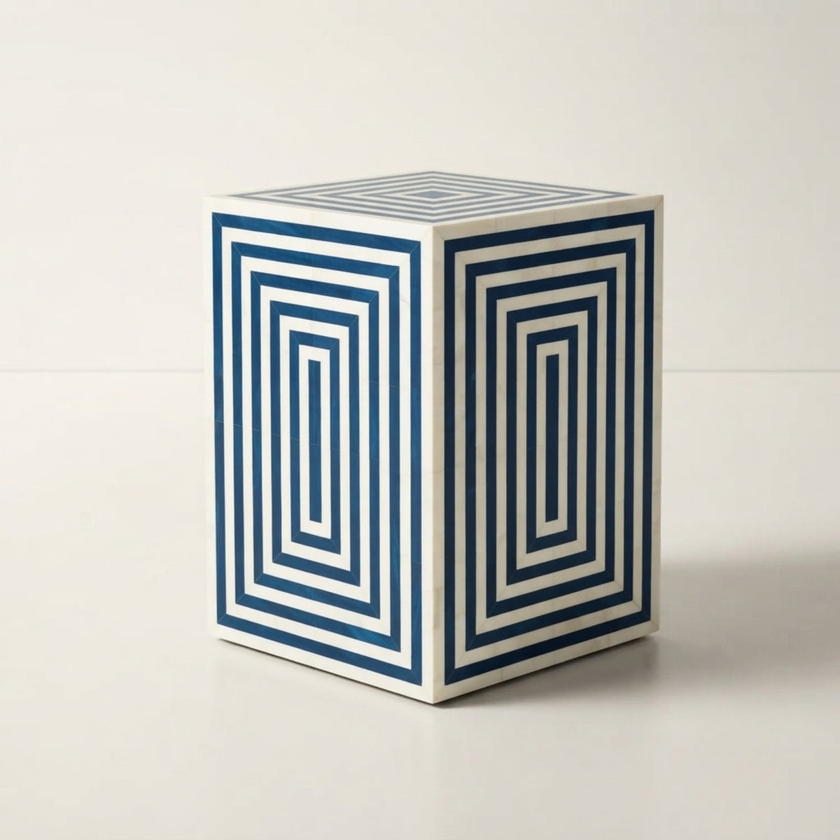 A cube-shaped side table or stool with a white base and navy blue geometric concentric rectangle patterns on two visible sides, photographed on a clean white studio surface.