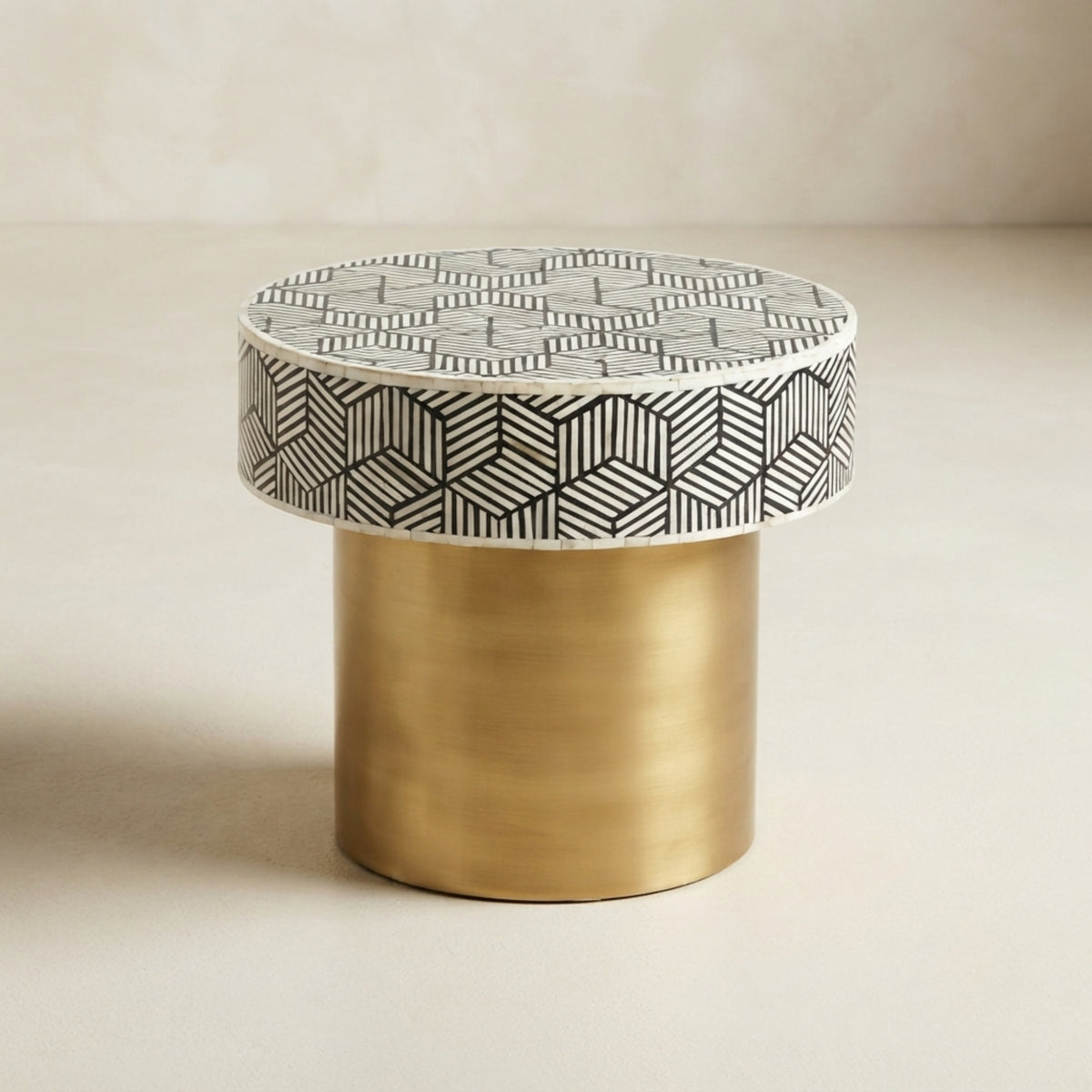 A modern cylindrical side table with a brushed gold metal base and a top upholstered in a black and white geometric patterned fabric, set against a neutral beige background.